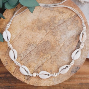 Può includere: Una collana di conchiglie bianche con perline di legno marrone naturale su uno stringa beige. La collana è esposta su una superficie di legno.