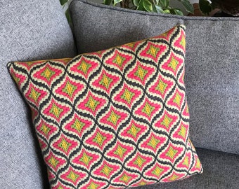 Puede incluir: Un cojín decorativo con un patrón geométrico en rosa, amarillo, negro y verde. El cojín está colocado sobre un sofá gris. El diseño del cojín presenta un motivo repetido en forma de diamante.