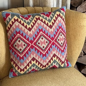Pode incluir: Uma almofada decorativa quadrada com um padrão geométrico em tons de vermelho, azul, rosa e amarelo. A almofada está colocada numa cadeira amarela. O padrão apresenta formas de diamante e ziguezague.