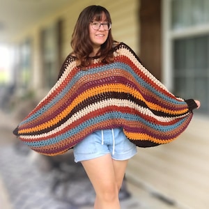 Puede incluir: Un poncho de ganchillo de corte holgado y manga larga con rayas horizontales en tonos marrón oscuro, crema, amarillo mostaza, burdeos y azul verdoso. La prenda presenta un escote ancho y un patrón de punto abierto con textura.
