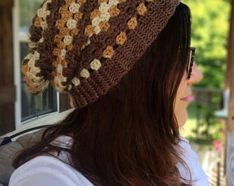 May include: A slouchy crochet beanie featuring a granny square pattern in alternating shades of dark brown, light brown, and cream. The hat has a solid dark brown ribbed brim and a relaxed, loose fit that hangs slightly toward the back of the head.