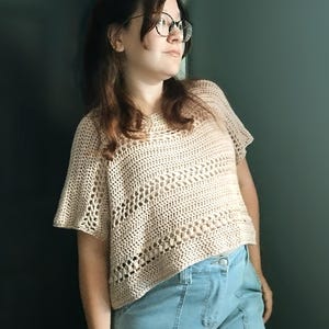 Puede incluir: Top de ganchillo beige de manga corta con un diseño de tejido suelto y abierto. El top tiene un cuello redondo y una forma ligeramente acampanada. El modelo lleva vaqueros azul claro.