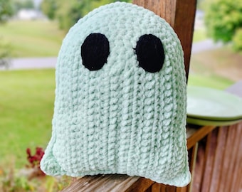 May include: A soft, mint green ghost-shaped cushion with two large, black eyes. The cushion is made from a textured material and is resting on a wooden railing. The background is blurred, showing green foliage.