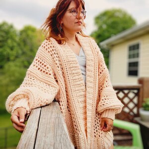 May include: A light pink crocheted cardigan with a loose fit and open front. The cardigan has a textured stitch pattern and is worn over a gray tank top.