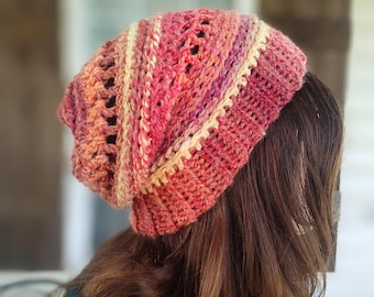 May include: A slouchy crochet beanie featuring a variegated yarn pattern in shades of red, orange, pink, and cream. The hat has a ribbed brim and a textured stitch pattern with openwork details, creating a layered appearance in warm, vibrant tones.
