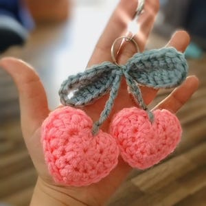 May include: A handmade crochet keychain featuring two pink heart-shaped cherries with gray stems and a gray leaf detail. The keychain has a silver ring for attaching to keys or bags. The soft, textured yarn gives a handcrafted feel.