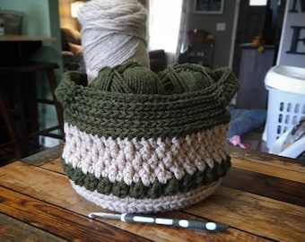 May include: A crocheted basket with a green and beige striped pattern. The basket is filled with balls of yarn in beige and green.