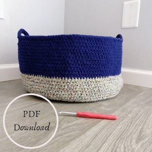 May include: A crocheted storage basket with a blue top and a multi-colored bottom. The basket is sitting on a wooden floor. A red crochet hook is lying on the floor next to the basket. The text "PDF Download" is in a circle on the floor.