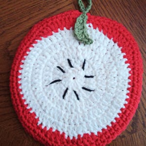 May include: A handmade crochet apple-shaped trivet. The trivet features a red border, a white center, and a green stem and leaf. The apple design includes black seed details. This kitchen accessory is designed to protect surfaces from hot cookware.
