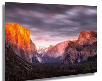 May include: A landscape photograph of a mountain valley at sunset. The towering rock formations are illuminated with golden light, contrasting with the dark forest below. A waterfall cascades down a cliff face, and the sky is filled with dramatic clouds.