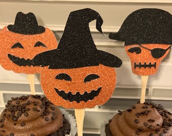 May include: Three chocolate cupcakes with brown frosting and dark chocolate sprinkles, each topped with a glittery orange and black Halloween-themed paper topper on a wooden stick, featuring a pumpkin with a hat, a witch hat, and a pirate skull.