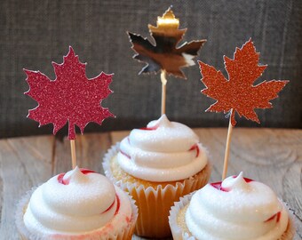 May include: Three cupcakes in white paper liners topped with white frosting and red swirls. Each cupcake features a decorative maple leaf topper on a wooden pick: one red glitter, one metallic silver, and one copper glitter maple leaf.