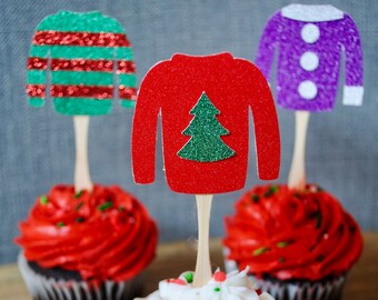 May include: Three cupcakes with festive sweater-shaped toppers. One cupcake has white frosting with sprinkles, the others have red frosting. The toppers are red, green, and purple with glitter accents, ideal for a holiday celebration.