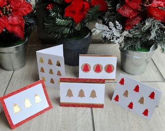 May include: A selection of Christmas cards with festive designs. Some cards have gold or red glitter borders, while others feature gold or red tree cutouts. The cards are displayed on a wooden surface, with poinsettias and greenery in the background.