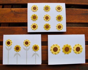 May include: Three white cards with yellow and brown paper sunflower cutouts. The top card has nine sunflowers, the middle card has four sunflowers, and the bottom card has three sunflowers.