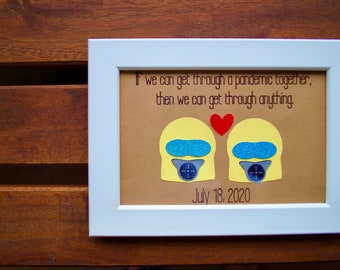 May include: A framed print with a brown background and white frame. The print features a quote about getting through a pandemic together, two yellow figures with blue and white scuba masks, and a red heart. The date July 18, 2020 is printed below the quote.