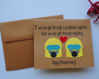 May include: A brown card with the text "If we can get through a pandemic together, then we can get through anything. Happy Anniversary!"  Two yellow paper cutouts of people wearing face masks with a red heart between them.