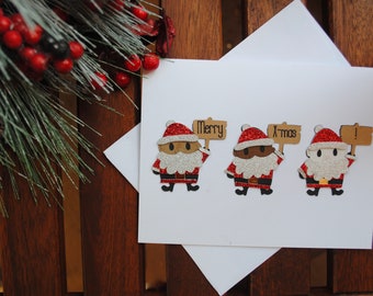 May include: A white Christmas card featuring three Santa Claus figures, each holding a sign. The signs read "Merry" and "Xmas". The Santas have red hats, black belts, and glittery beards. The card is on a wooden surface.