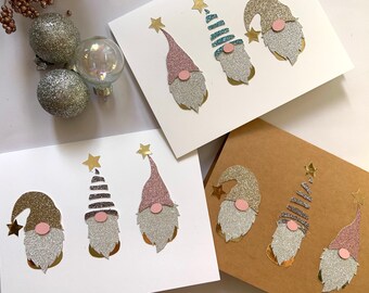 May include: Handmade Christmas cards with glittery gnome designs in pink, silver, and gold. Each card features a unique gnome with a star-topped hat and a round pink nose. The cards are on white and brown cardstock.