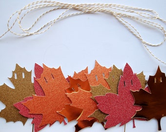 May include: A collection of die-cut maple leaf shapes in shades of gold, copper, and red, featuring both glittery and metallic finishes. Each leaf has two small holes at the top for stringing, accompanied by a length of thin, twisted beige twine.