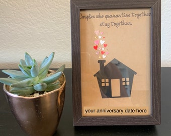 May include: A framed print with a brown background and a black silhouette of a house with a chimney. The chimney has a cloud of pink and red hearts coming out of it. The text "Couples who quarantine together stay together." is written in black cursive. The text "your anniversary date here" is written in black below the house.
