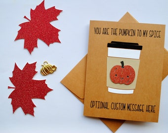 May include: A tan greeting card featuring a paper cutout of a coffee cup with a glittery orange pumpkin, black text reading YOU ARE THE PUMPKIN TO MY SPICE and OPTIONAL CUSTOM MESSAGE HERE, accompanied by two red glittery maple leaves and a gold pumpkin charm.