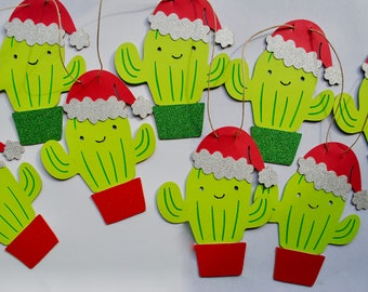 May include: A selection of Christmas cactus decorations. Each decoration is a light green cactus with a smiling face, a red pot, and a red Santa hat with silver glitter trim. They are strung with twine.