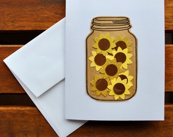May include: A white card featuring a brown mason jar filled with yellow and brown glitter sunflowers. The card is on a wooden surface.
