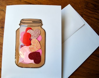 May include: A white card with a brown paper mason jar filled with pink, red, and gold glitter hearts. The jar is glued to the card.