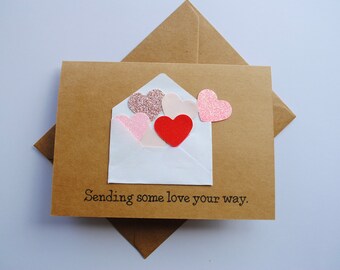 May include: A brown card with a white envelope containing pink and red glitter hearts. The card reads "Sending some love your way."
