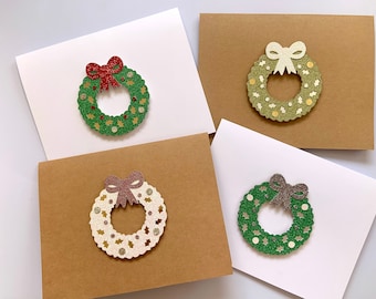 May include: Four Christmas cards with glittery wreath decorations. Two cards are white, and two are brown. The wreaths are green, gold, and white, with red, gold, and brown bows. The wreaths have star and dot accents.