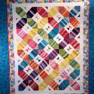 May include: A colorful handmade quilt with a patchwork design. The quilt features a variety of fabrics in shades of red, orange, yellow, green, blue, and purple. The quilt has a white border and a floral patterned edge. The quilt is displayed on a blue background.
