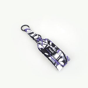 Puede incluir: Una correa de mu&ntilde;eca para llavero estampada con un anillo de metal negro. La correa presenta un dise&ntilde;o de formas moradas, negras, blancas y grises. La palabra "hosh" est&aacute; impresa en una etiqueta negra en la correa.