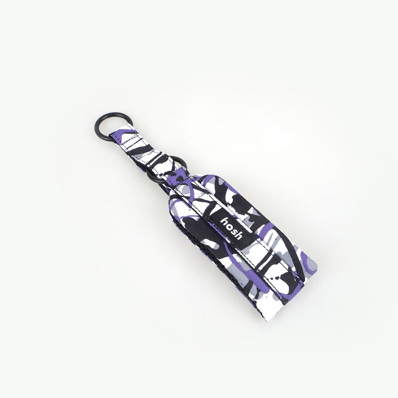 Puede incluir: Una correa de mu&ntilde;eca para llavero estampada con un anillo de metal negro. La correa presenta un dise&ntilde;o de formas moradas, negras, blancas y grises. La palabra "hosh" est&aacute; impresa en una etiqueta negra en la correa.