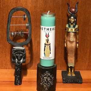Op de afbeelding: Een turquoise kaars met een wit label waarop "Hetheru Neteru" staat en een zwart-gouden afbeelding van een Egyptische godin. Het label bevat ook de tekst "Purified Sight & Divine Beauty". De kaars staat op een zwarte standaard met gouden accenten. Links staat een zwart houten muziekinstrument met metalen ringen en een gegraveerd gezicht. Rechts staat een gouden beeld van een Egyptische godin met een gouden kroon en een gouden jurk.