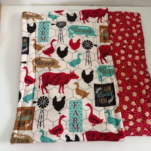 May include: A rectangular quilted fabric mat featuring a cream background with a black wire fence pattern. The print includes red cows, black chickens, teal geese, and black windmills. Text reads Farm Fresh Everyday and Farm Sweet Farm. The reverse is red with white daisies.