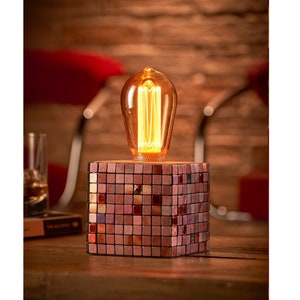 May include: A small table lamp with a mosaic base made of small, square tiles in shades of pink, red, and brown. The lamp has a clear glass bulb with a warm, incandescent light.