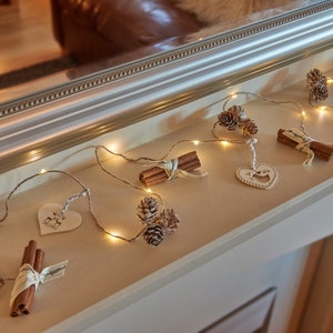 May include: A string of warm white fairy lights with cinnamon sticks, pine cones, and a white ceramic heart with the text "Merry Christmas" hanging from it.