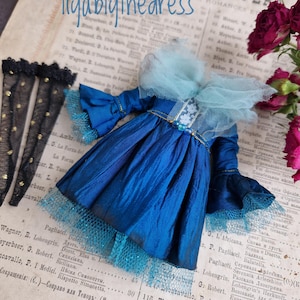 Op de afbeelding: Een miniatuur blauwe poppenjurk met een geplooide rok en ruchesmouwen. De jurk heeft een lichtblauwe tule kraag en bies. Zwarte kanten kousen met gouden stippen zijn links zichtbaar. De tekst "liyablythedress" staat bovenaan.