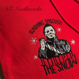 May include: Red hooded sweatshirt with an embroidered design featuring a masked figure holding a knife, snowflakes, and the text "SLASING THROUGH THE SNOW". The brand "NE Needleworks" is embroidered at the top.