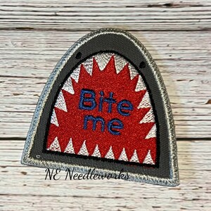 May include: A gray felt patch with a white and red embroidered shark mouth with the text "Bite me" in blue thread. The patch is stitched with a white border and has the text "NE Needleworks" in black thread at the bottom.