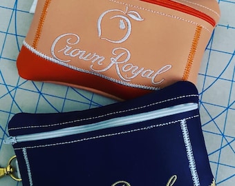 Op de afbeelding: Twee ritszakjes met Crown Royal branding. Eén zakje is perzikkleurig met witte borduursels en een perzikontwerp. Het andere zakje is donkerblauw met gouden borduursels.