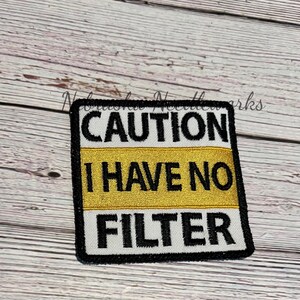 May include: Embroidered patch with the text "CAUTION I HAVE NO FILTER" in black, white, and gold. The patch is square with a black border and is likely designed to be sewn or ironed onto clothing or accessories.