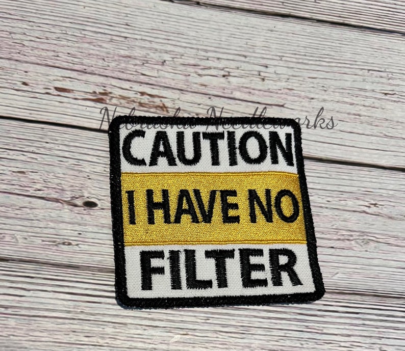 May include: Embroidered patch with the text "CAUTION I HAVE NO FILTER" in black, white, and gold. The patch is square with a black border and is likely designed to be sewn or ironed onto clothing or accessories.
