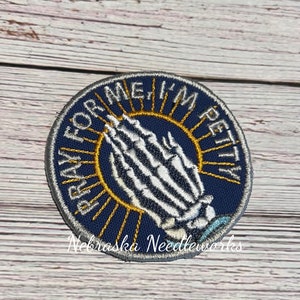 May include: A blue and white embroidered patch with a skeleton hand in a praying position. The text around the hand reads "Pray for me I'm petty".