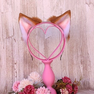May include: A pair of faux fur cat ears with a pink heart-shaped headband. The headband is on a pink stand with pink flowers.