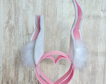 May include: A pink bunny ear headband with a heart-shaped design, featuring white and pink ears with fluffy white accents. The headband is displayed with pink and white flowers.