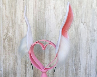 May include: White bunny ears with pink lining on a pink heart-shaped stand with a floral arrangement at the base.