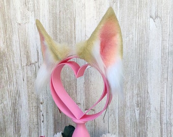 May include: A pair of faux fur cat ears with a pink and white ombre design. The ears are attached to a headband and are displayed on a pink heart-shaped stand.