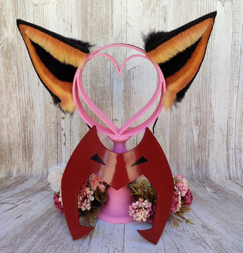 May include: A pair of orange and black furry cat ears with a pink heart-shaped headband. The headband is attached to a red plastic mask with a black eye design.